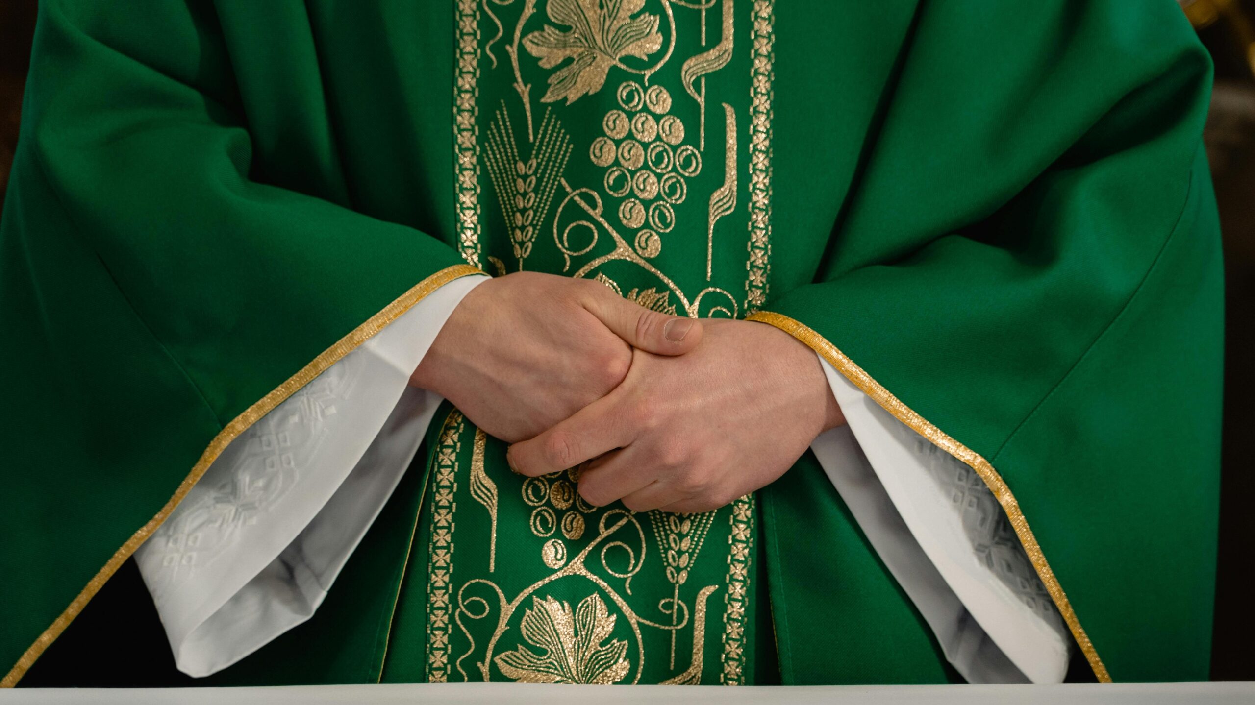 A close-up view of a priest's hands clasped in prayer wearing a green chasuble with gold embroidery.